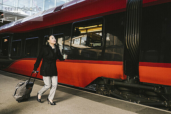 Geschäftsfrau mit Gepäck  die ein Getränk in der Hand hält  während sie auf dem Bahnhof durch den Zug läuft
