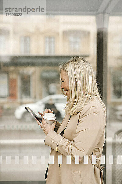 Seitenansicht einer lächelnden Geschäftsfrau  die an einer Bushaltestelle ein Smartphone benutzt