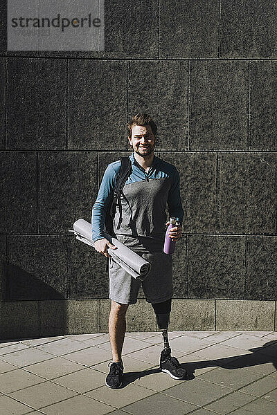 Porträt eines lächelnden Mannes mit Behinderung  der eine Wasserflasche und eine Gymnastikmatte auf einem Fußweg an einem sonnigen Tag hält