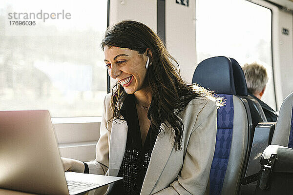 Glückliche Geschäftsfrau mit In-Ear-Kopfhörern bei einem Videogespräch über einen Laptop im Zug