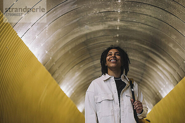 Glückliche junge nicht-binäre Person mit Rucksack in beleuchtetem Tunnel stehend