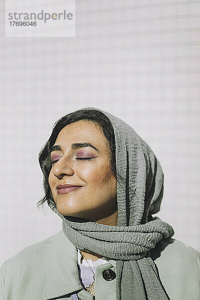 Lächelnde Frau mit Kopftuch steht mit geschlossenen Augen an einem sonnigen Tag