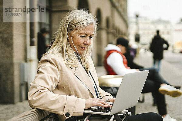 Blonde Geschäftsfrau  die einen Laptop benutzt und auf einer Bank in der Stadt sitzt