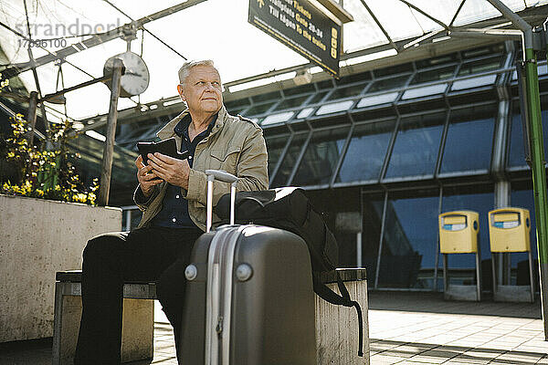 Geschäftsmann  der wegschaut und sein Smartphone in der Hand hält  während er auf einer Bank mit Gepäckstücken am Bahnhof sitzt