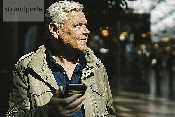 Lächelnder älterer Geschäftsmann  der am Bahnhof über sein Smartphone nachdenkt