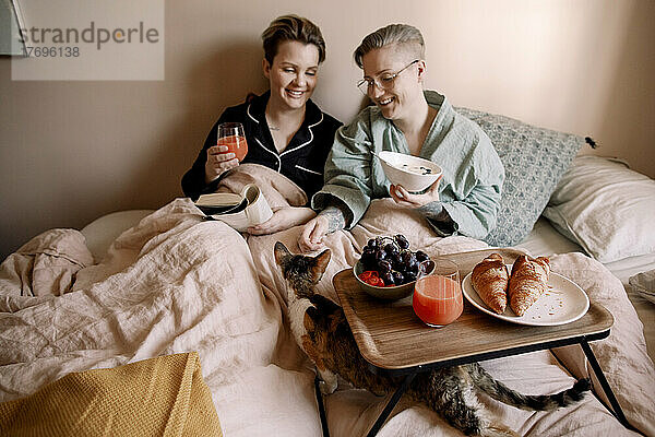 Lächelndes lesbisches Paar beim Frühstück im Bett mit Katze zu Hause