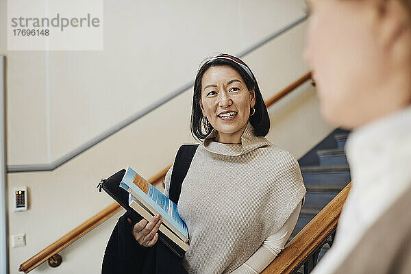 Lächelnde Lehrerin mit Büchern in der Hand und Blick auf einen Kollegen in der Universität