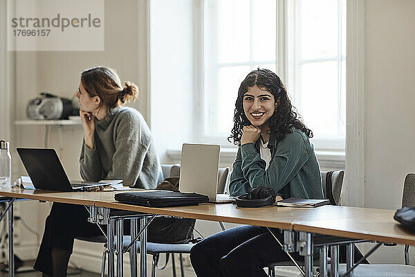 Porträt einer lächelnden Studentin mit Laptop  die mit einem Freund am Schreibtisch im Klassenzimmer sitzt