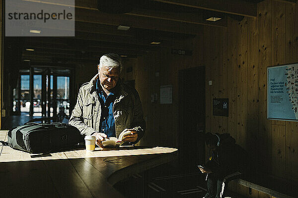 Ein älterer Geschäftsmann liest ein Buch  während er im Wartezimmer eines Bahnhofs in der Sonne steht.