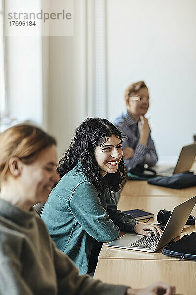 Glückliche Studentin mit Laptop  die inmitten von Freunden am Schreibtisch im Klassenzimmer sitzt