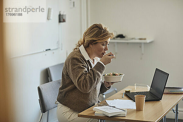 Professorin isst zu Mittag  während sie am Schreibtisch mit Laptop im Klassenzimmer sitzt