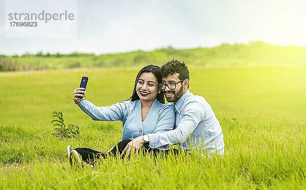 Lächelndes verliebtes Paar  das im Gras sitzt und Selfies macht  Junges verliebtes Paar  das ein Selfie auf dem Feld macht  Verliebte  die auf dem Feld Selfies mit ihrem Smartphone machen