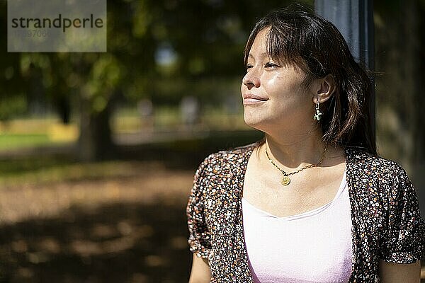 Porträt einer jungen lateinamerikanischen Frau in einem Park  die sich an einen Laternenpfahl lehnt und die Sonnenstrahlen auf ihrer Haut genießt