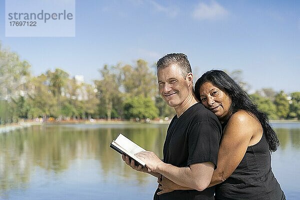 Älteres multiethnisches Paar  das sich an einem See umarmt und ein Buch liest  lächelnd in die Kamera schaut. Raum kopieren