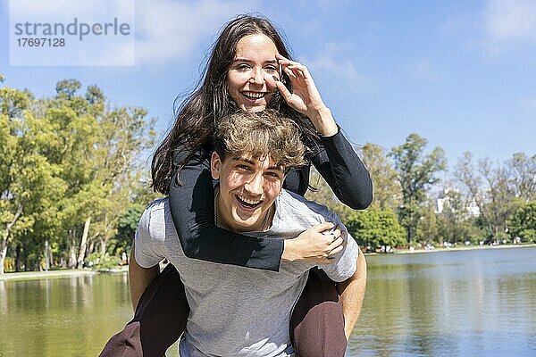 Junges Paar an einem See. Freund trägt seine Freundin auf dem Huckepack. Lustiger und glücklicher Ausdruck