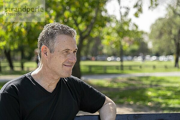 Nahaufnahme eines reifen Mannes mit grauem Haar  der ein schwarzes T-Shirt trägt und auf einer Parkbank sitzt und zur Seite schaut