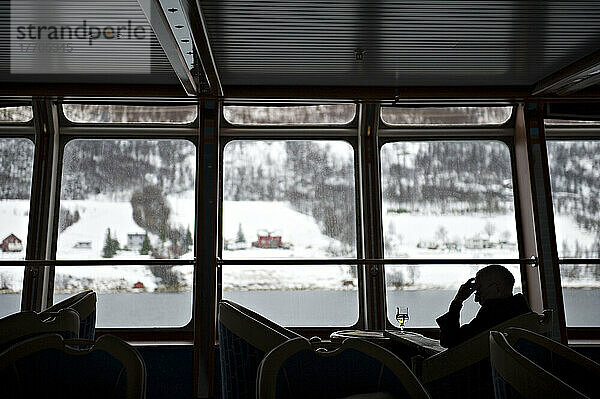 Ein Passagier liest die Zeitung in der Panoramalounge des Hurtigrutenschiffs Frau Nordlys  während draußen die schneebedeckte Landschaft vorbeizieht; Norwegen