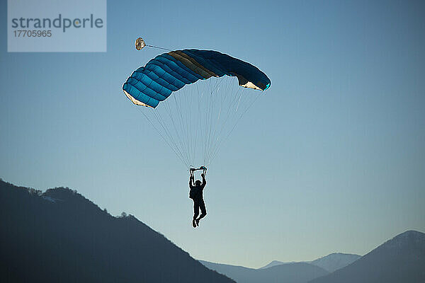 Ein Fallschirmspringer schwebt mit einem Fallschirm gegen einen blauen Himmel und die Schweizer Alpen; Locarno  Tessin  Schweiz