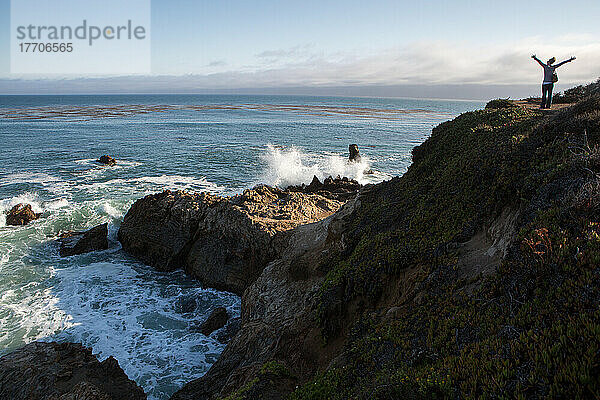 Eine Touristin auf einer Felsformation am Pazifischen Ozean. Als die Wellen an die Felsen schlagen  streckt sie begeistert ihre Arme aus; San Simeon Cove  San Simeon  Kalifornien