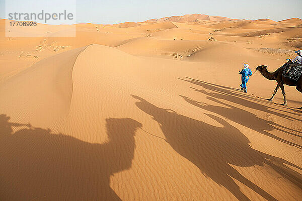 Ein Mann führt einen Kamelzug an  während Schatten von Kamelen und Menschen die Sanddünenlandschaft bedecken; Erg Chebbi  Sahara-Wüste  Marokko