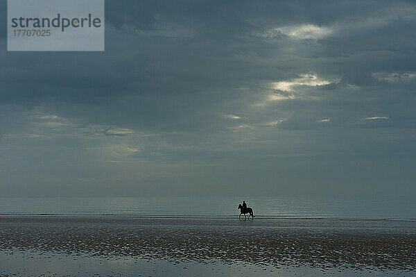 Person Reiten Pferd entlang Camber Sands auf bewölktem Tag mit Sonnenstrahl; East Sussex  England