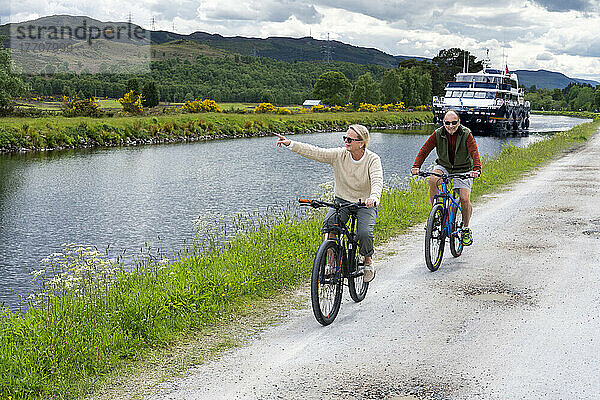 Ein Mann und eine Frau fahren mit dem Fahrrad auf dem Treidelpfad des Caledonian Canal in der Nähe von Fort Augustus  Schottland. Im Hintergrund fährt ein Ausflugsboot den Kanal entlang; Fort Augustus  Schottland