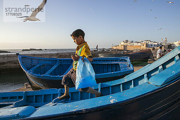 Ein kleiner Junge überquert behutsam ein Boot am Ufer von Essaouira  Marokko; Essaouira  Marokko