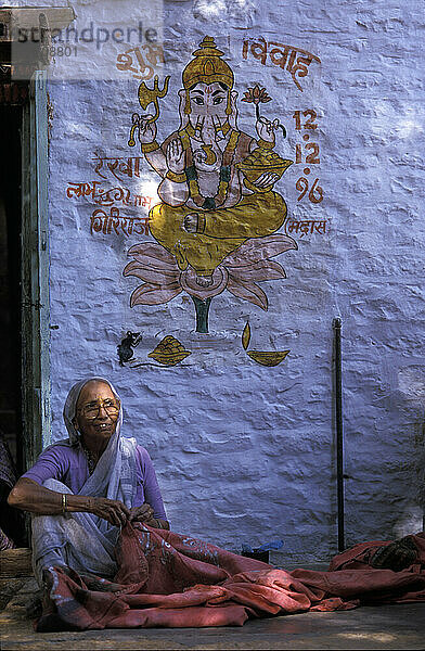 Nähende Frau vor blauer Wand mit Ganesh-Gemälde; Jaisalmer  Rajasthan  Indien