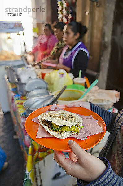 Frauenhände halten einen Teller mit einheimischem mexikanischem Essen  eine Gordita  neben dem Stand eines Lebensmittelverkäufers; San Luis De La Paz  Guanajuato  Mexiko