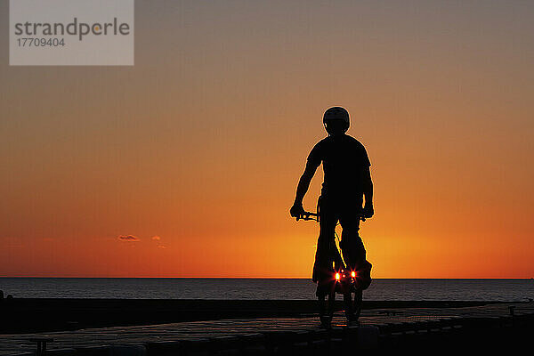 Silhouette eines jungen Mannes auf einem Stuntfahrrad im BMX-Stil  der bei Sonnenuntergang an der West Bay  an der Jurassic Coast  South West Coast Path  Dorset  England  Fahrradstunts und -tricks vorführt