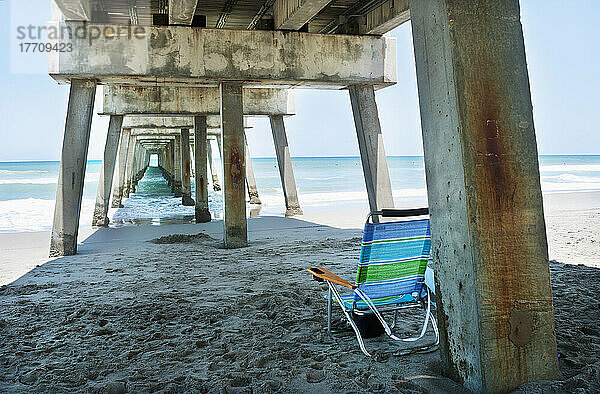 Ein blau  grün und weiß gestreifter Strandkorb sitzt leer unter einem Pier am Jupiter Beach; Florida  Vereinigte Staaten von Amerika