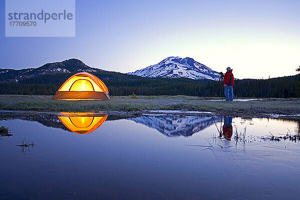 Mann mit Kamera und Stativ fotografiert die Schönheit Oregons bei Sonnenaufgang neben einem Zelt an einem Teich  mit South Sister und Broken Top im Hintergrund  Oregon Cascades; Oregon  Vereinigte Staaten von Amerika