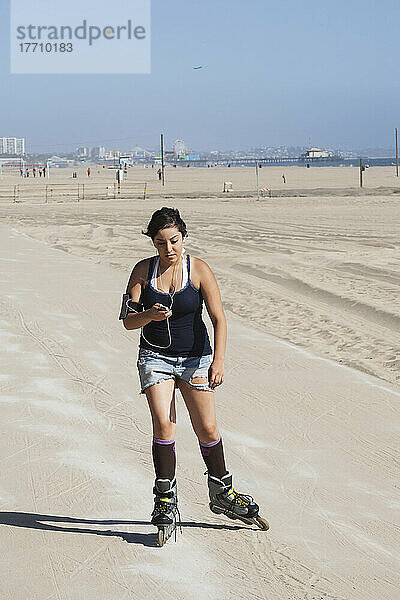 Eine junge Frau mit Inline-Skates auf der Promenade entlang der Küste mit Ohrstöpseln und einem Mobiltelefon; Kalifornien  Vereinigte Staaten von Amerika