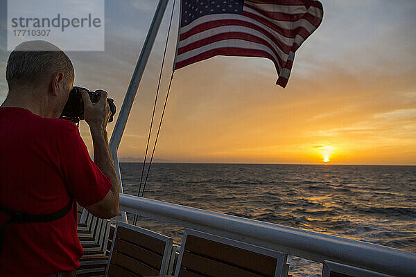 Inmitten eines farbenprächtigen Morgenhimmels in der Nähe von Panama fotografiert ein Passagier an Bord eines Expeditionsschiffes den Sonnenaufgang  während eine amerikanische Flagge im Wind weht; Panama