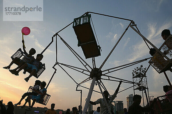 Silhouette von Kind mit Ballon und Kinder auf Fair Ground Ride bei Sonnenuntergang am Chowpatty Strand  neue Entwicklung auf Malabar Hill im Hintergrund; Mumbai  Maharashtra State  Indien