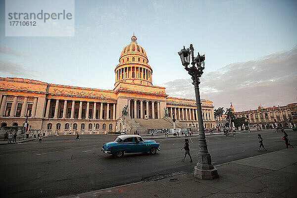 Menschen gehen durch die Straßen und ein amerikanischer Oldtimer fährt am El Capitolio-Gebäude in der Innenstadt von Havanna vorbei; Havanna  Kuba