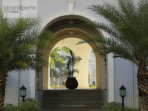 Eine Treppe führt zu einem gewölbten Eingang mit Laternenpfählen und Palmen