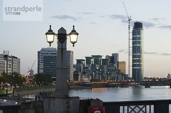 In der Dämmerung beleuchteter Laternenpfahl und Gebäude entlang der Waterfront; London  England
