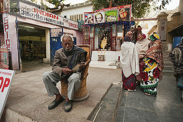 Menschen stehen im Gespräch und ein Mann sitzt in einem Stuhl außerhalb von Einzelhandelsgeschäften; Udaipur  Indien