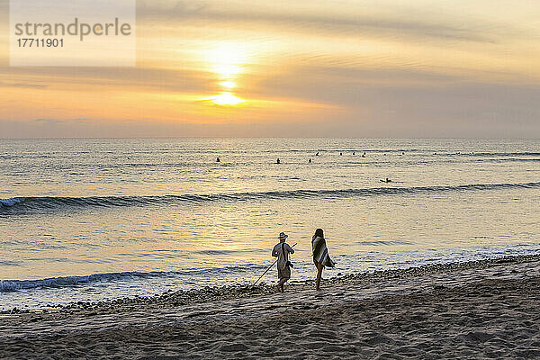 Surfer im Wasser und Menschen  die bei Sonnenuntergang am Strand spazieren gehen  San Onofre State Beach; San Clemente  Kalifornien  Vereinigte Staaten