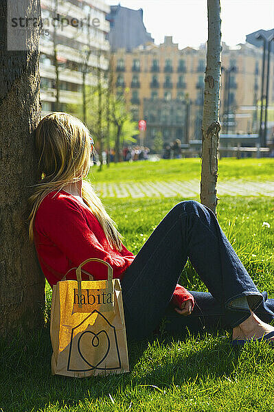Eine junge Frau sitzt mit einer Einkaufstasche gegen einen Baum auf dem Rasen; Barcelona  Spanien
