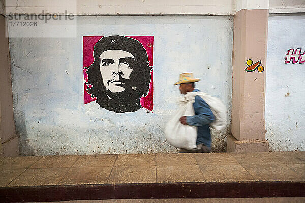 Ein Mann trägt eine Last neben einem Bild von Che Guevara an der Wand eines kubanischen Bauernmarktes; Cienfuegos  Kuba