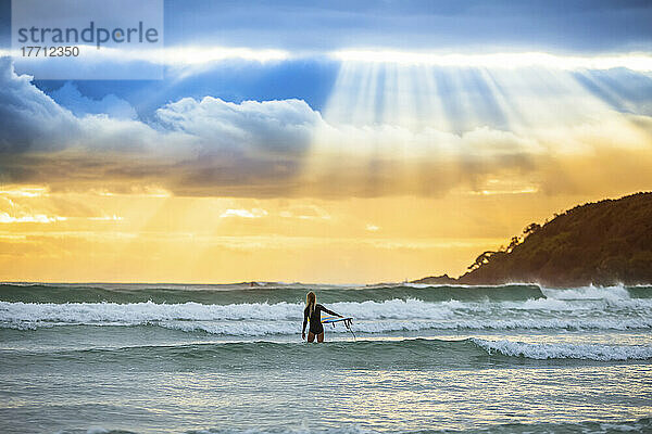 Ein wunderschöner Sonnenaufgang begrüßt eine Surferin  die sich auf das Surfen am frühen Morgen vorbereitet und hinauspaddelt; Arrawarra  New South Wales  Australien