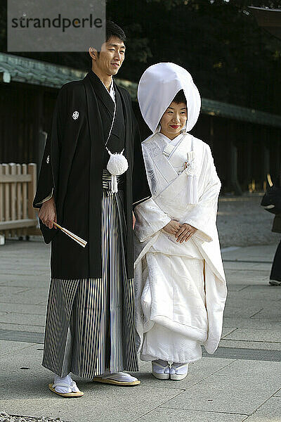 Nach der traditionellen japanischen Shinto-Hochzeitszeremonie versammeln sich die Familien zu einem formellen Porträtshooting am Meiji-Jingu-Schrein in der Nähe des Bahnhofs Harajuku in Tokio. Hier posieren die Braut und der Bräutigam für eine informellere Aufnahme nach dem formellen Fototermin  bei dem mehrere Schichten des weißen Kimonos der Braut mit einem Seidenbrokat bedeckt sind. Traditionell ist die Braut von Kopf bis Fuß in Weiß gekleidet  als Symbol der Reinheit. Eine traditionelle Kopfbedeckung  Tsuno Kakushi genannt  was wörtlich übersetzt die Hörner verbergen bedeutet  wird ebenfalls von der Braut getragen  um zu zeigen  dass sie eine ruhige und geduldige Ehefrau sein wird. Das Ensemble des Bräutigams ist einfacher: Bei einer traditionellen Shinto-Hochzeitszeremonie trägt er eine dunkle Plisseehose und einen kurzen schwarzen Kimono mit dem Familiensiegel.