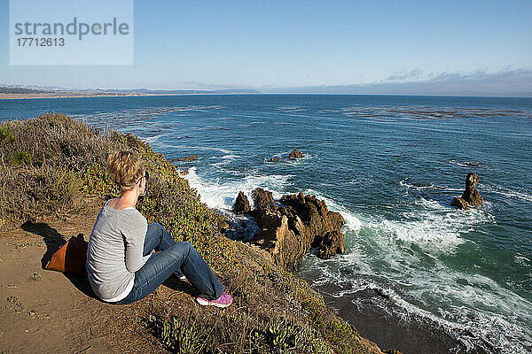 Ein Tourist sitzt an einem Aussichtspunkt am Pazifischen Ozean und sieht zu  wie die Wellen auf die Felsformationen prallen; San Simeon Cove  San Simeon  Kalifornien