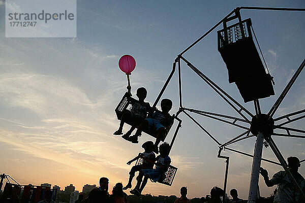 Silhouette von Kind mit Ballon und Kinder auf Fair Ground Ride bei Sonnenuntergang am Chowpatty Strand  neue Entwicklung auf Malabar Hill im Hintergrund; Mumbai  Maharashtra State  Indien