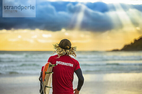 Blick von hinten auf einen Surflehrer  der auf den Ozean hinausblickt und von einem wunderschönen Sonnenaufgang begrüßt wird  während er sich auf eine frühmorgendliche Surfpartie vorbereitet; Arrawarra  New South Wales  Australien