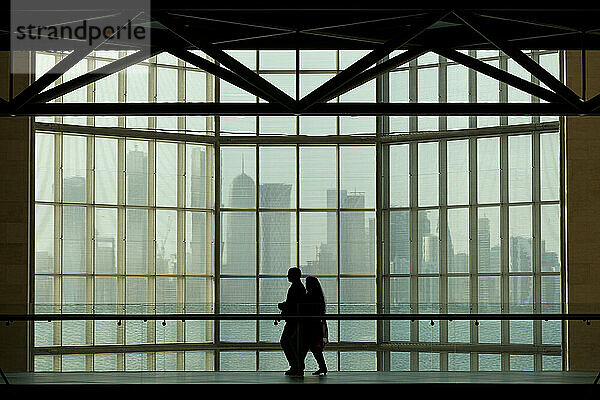 Silhouette eines Paares  das an einem großen Fenster im Museum für islamische Kunst vor der modernen Stadtsilhouette von Doha vorbeigeht; Doha  Katar