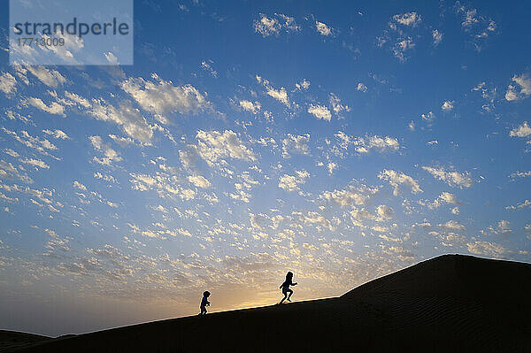 Zwei junge Mädchen laufen in der Abenddämmerung eine Sanddüne hinauf; Dubai  Vereinigte Arabische Emirate