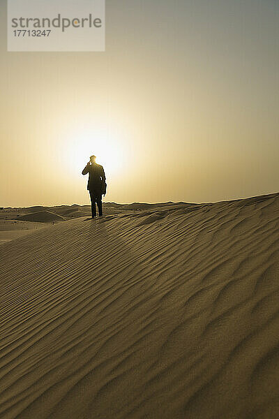 Man In Smart Suit Making Phone Call On Top Of Sand Dune At Dusk; Dubai  Vereinigte Arabische Emirate